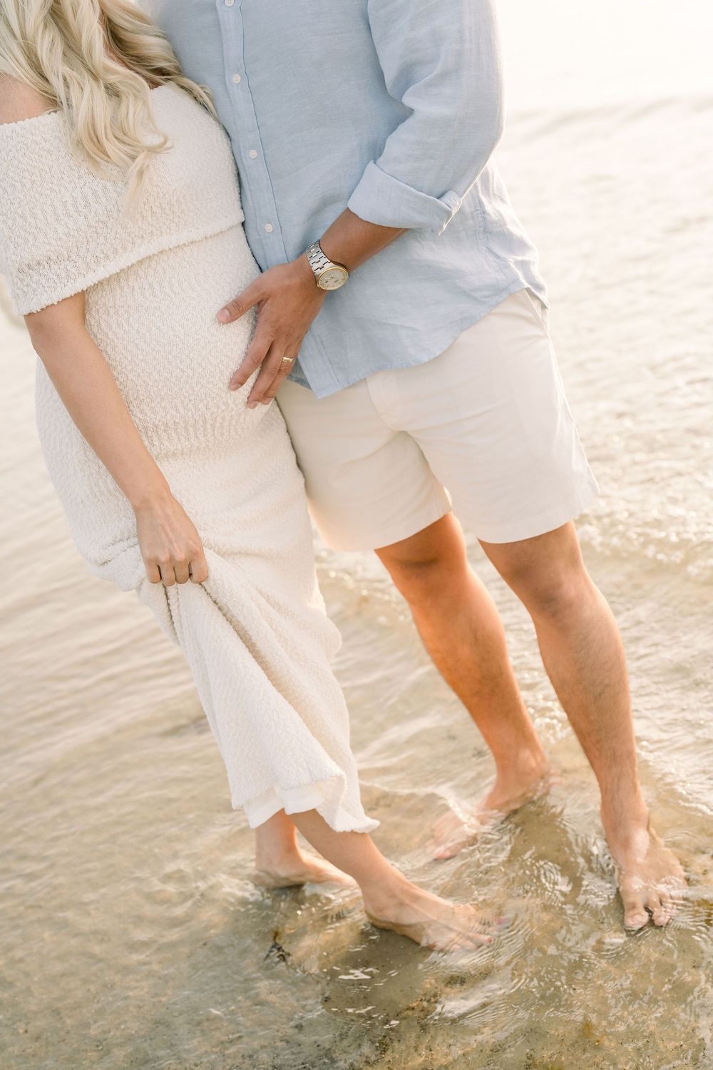Couple's feet splashing in shallow beach water wearing white and light blue casual attire.