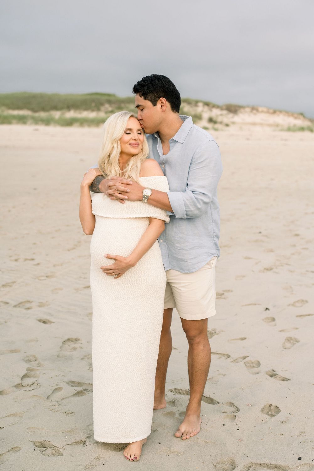 A couple shares intimate moments on a sandy beach wearing light colored outfits during a romantic photoshoot.