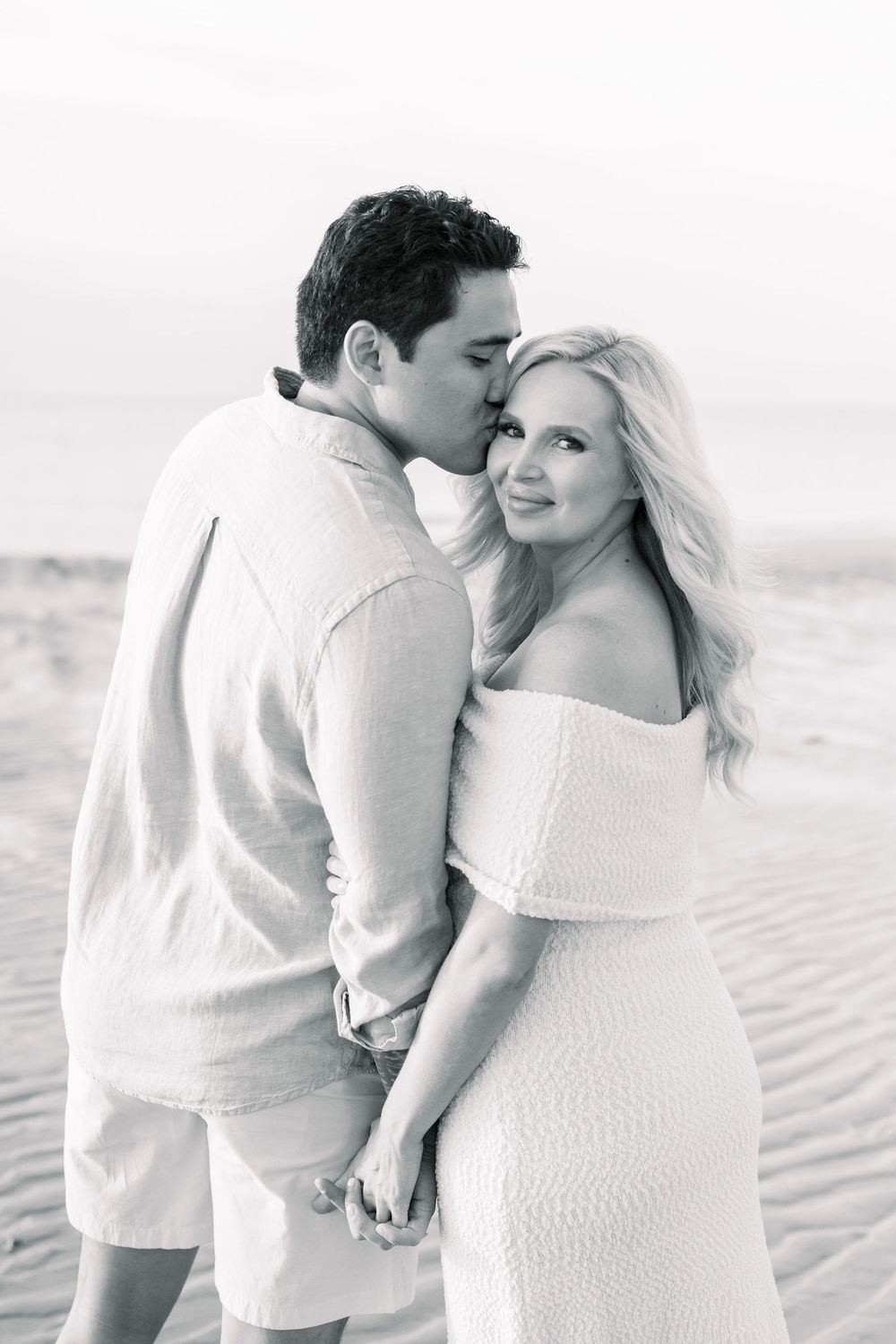 A romantic black and white beach photoshoot featuring a couple in white attire embracing on sandy dunes.