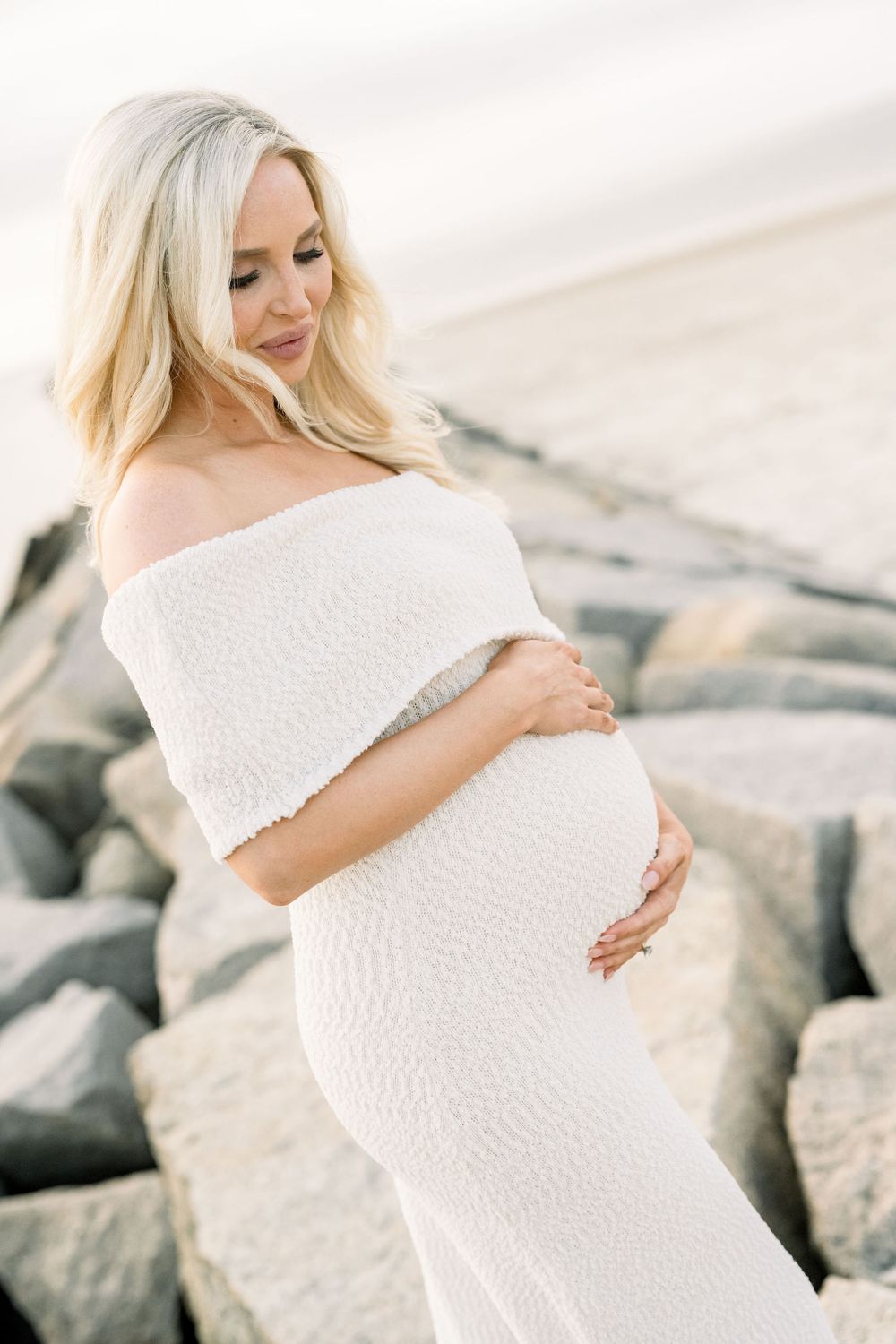 Expecting mother in flowing white dress poses on rocky beach during maternity photoshoot.