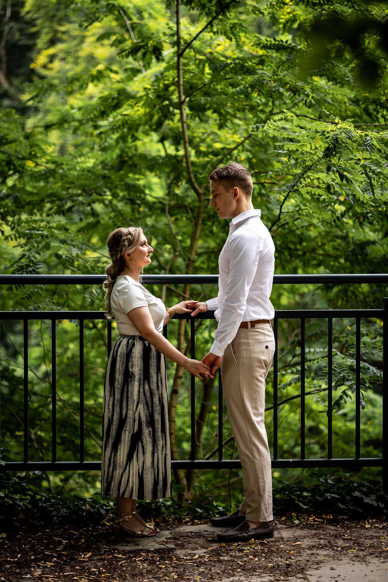A romantic couple stands against a railing surrounded by lush green foliage in a park setting.
