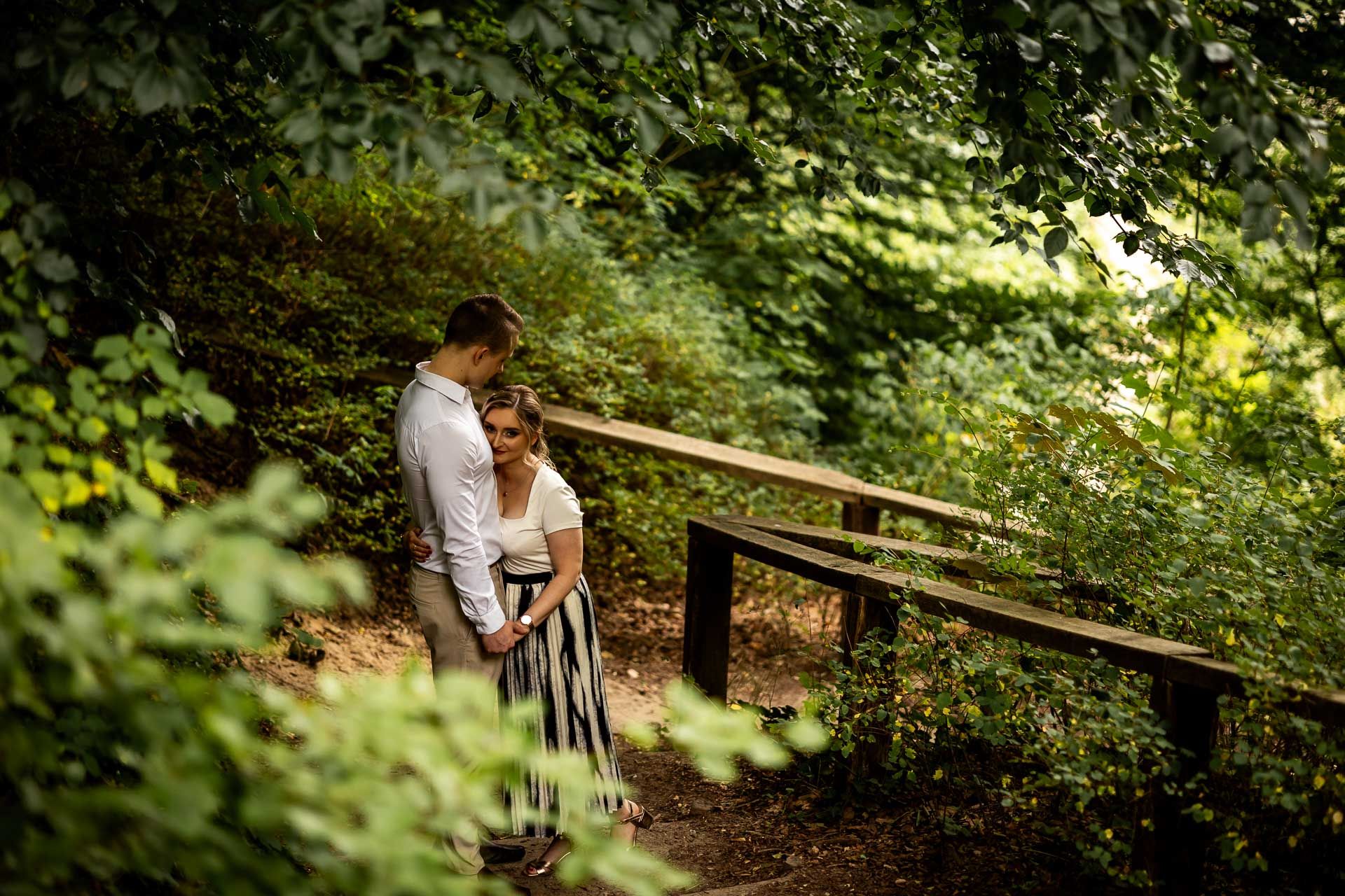 Couple photographed through tree branches on a wooded path with natural lighting.