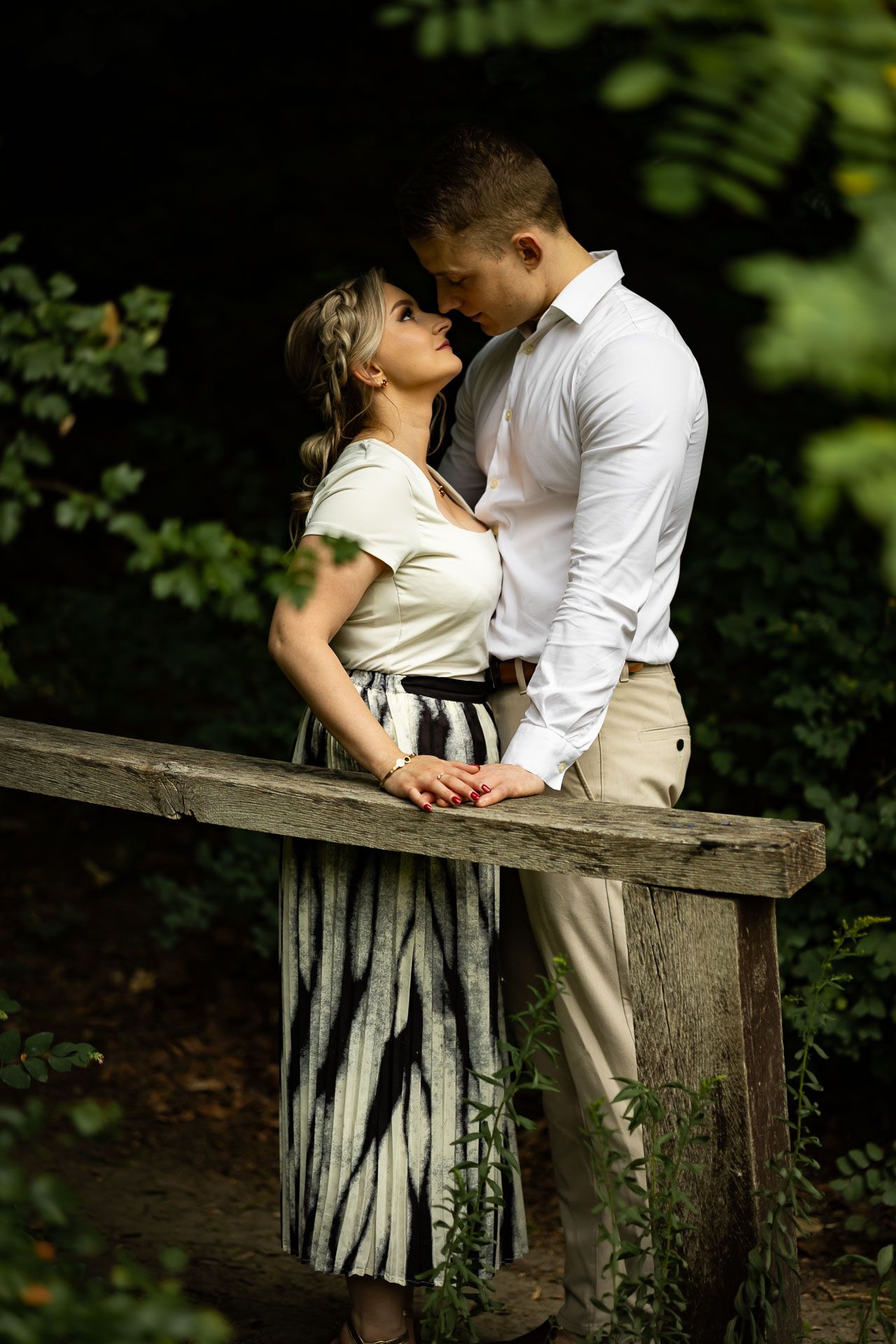 Romantic couple leaning against a wooden railing in a forest setting with dappled sunlight.