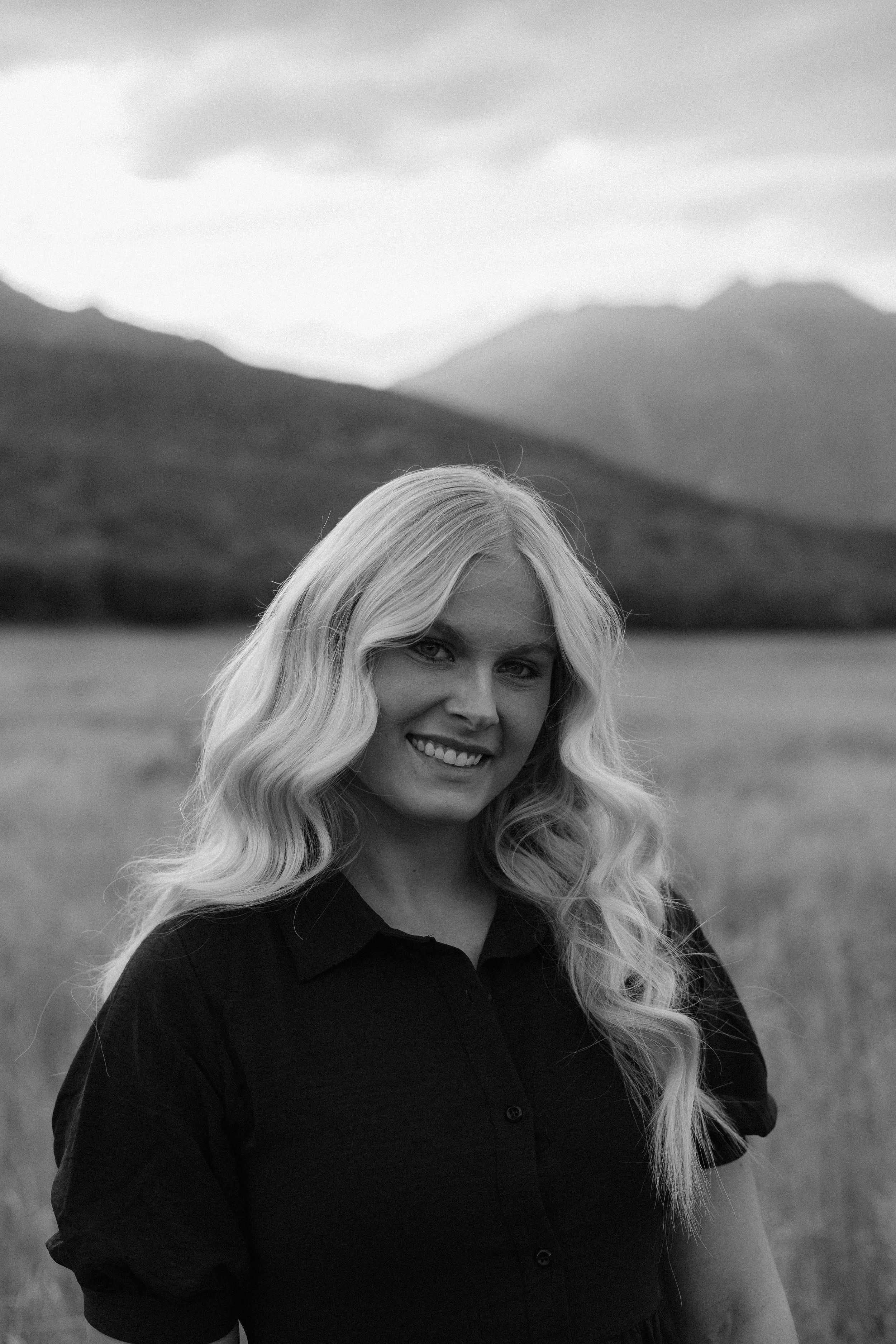 Person in black sweater smiles while sitting in mountain meadow with peaks in background.