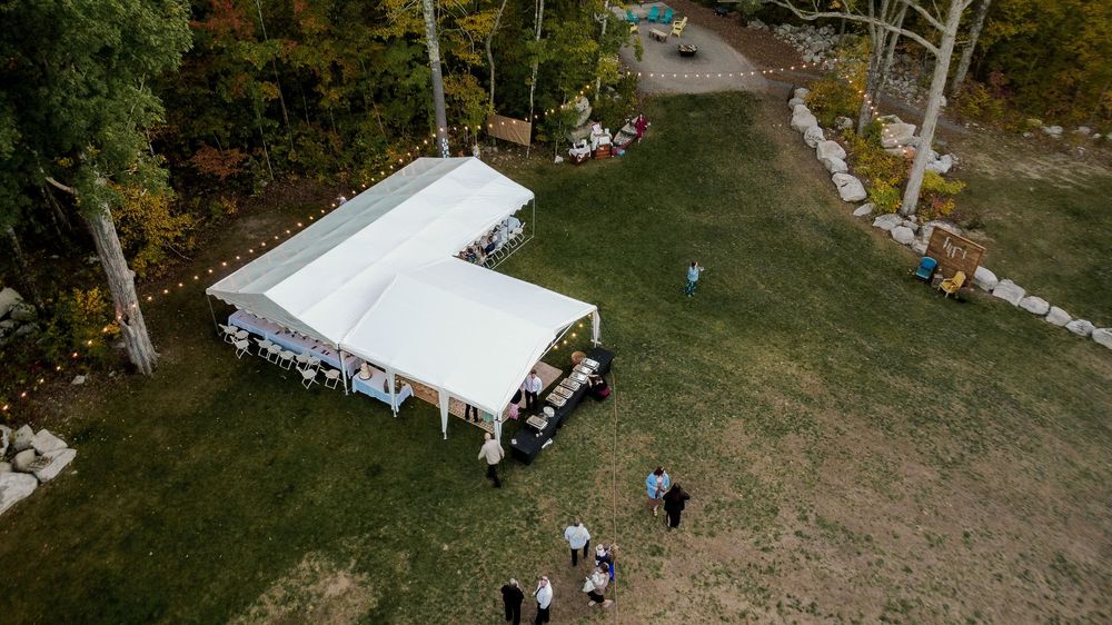 Aerial view of a white outdoor event tent set up on a grassy lawn with trees and people gathering underneath.