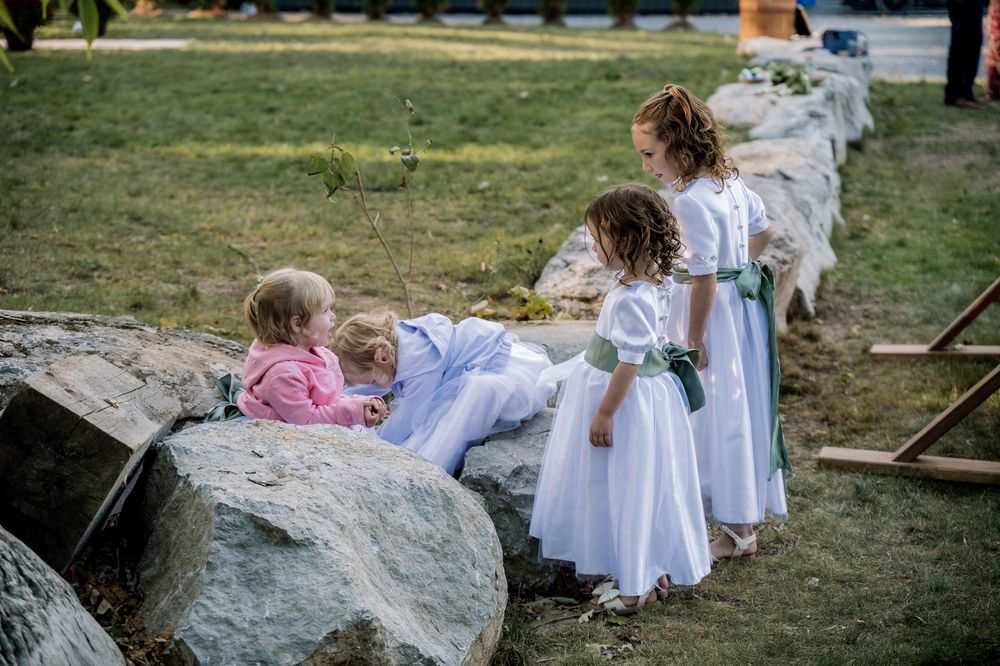 Children in white dresses explore a natural outdoor setting with rocks and grass on a sunny day.