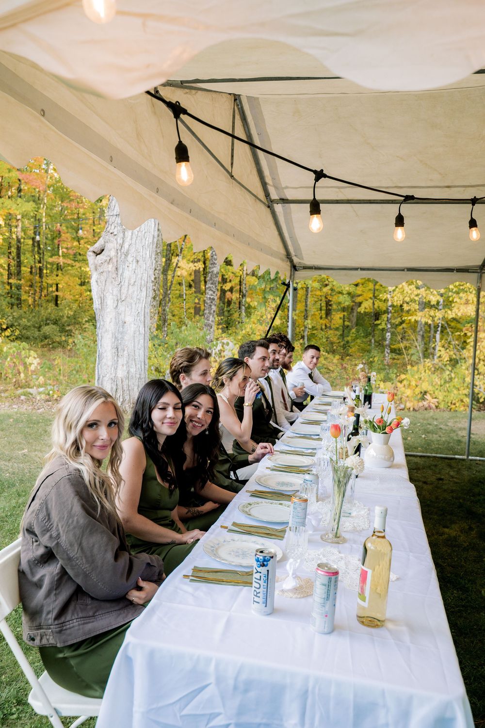 Long dining table set under a white tent canopy with guests seated outdoors in a wooded setting during daytime.