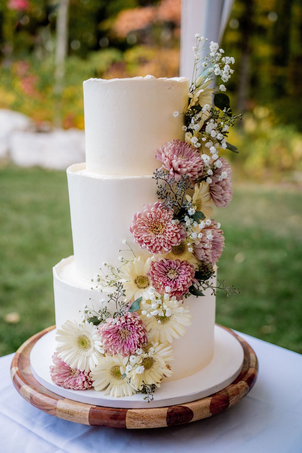 Elegant white wedding cake decorated with fresh pink and white flowers on wooden cake stand.