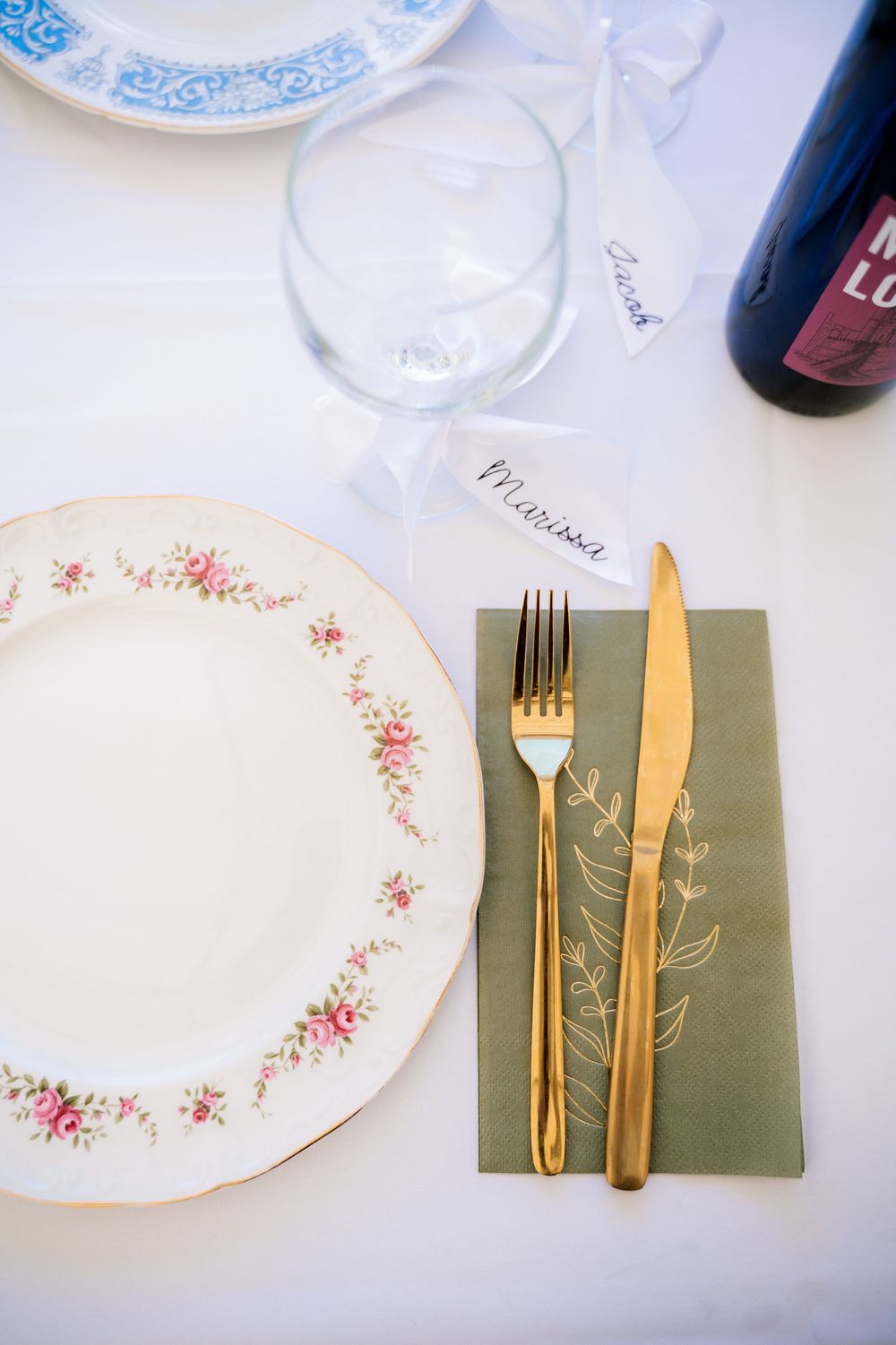 Elegant table setting with white floral plates and gold utensils on green napkins arranged in a formal dining setup.