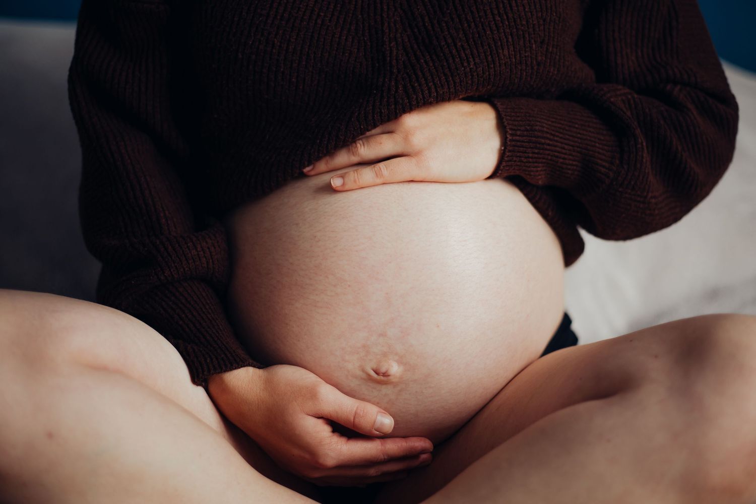 Pregnant woman in dark sweater cradling her bare baby bump with both hands while seated.