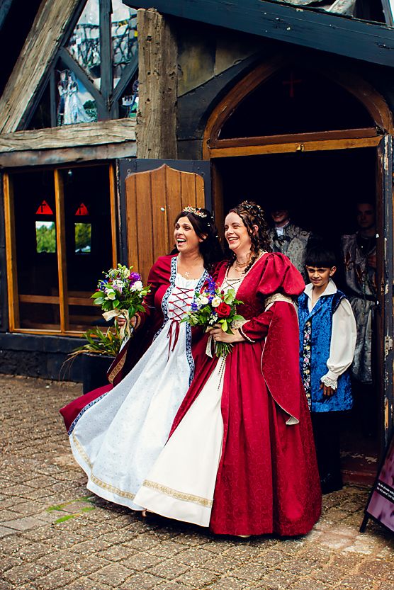 A group in historical period dress including red gowns and white dresses pose together outside a rustic wooden building.