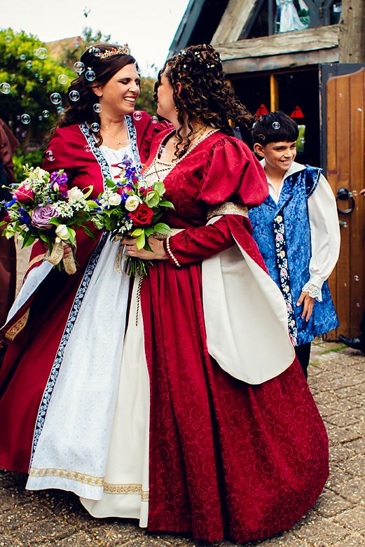 People in medieval renaissance costumes with red cloaks and white dresses pose together with floral bouquets.