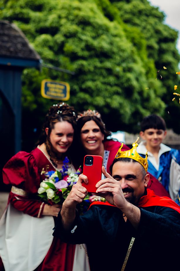 People in medieval costumes celebrate at a renaissance fair with someone taking photos on a smartphone.