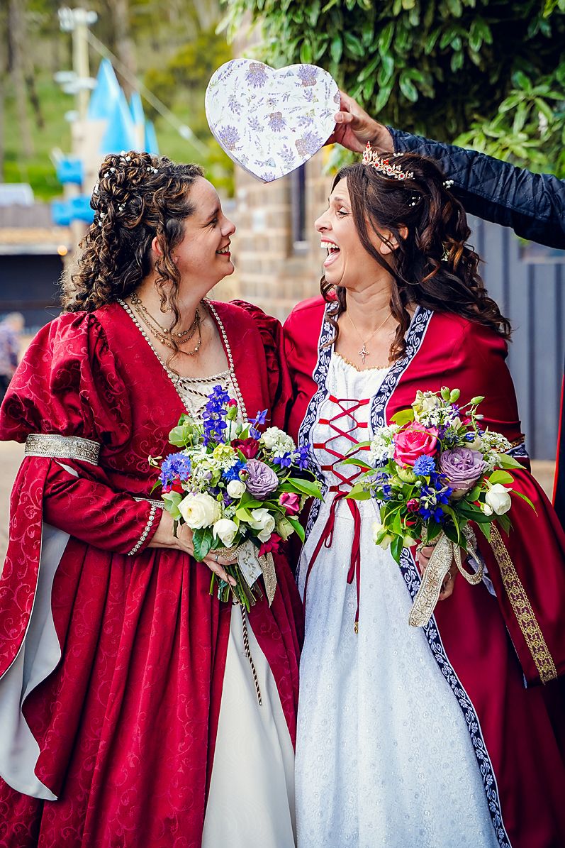 Two people in medieval renaissance faire costumes, one in red velvet and one in white, share joyful moments with flowers.