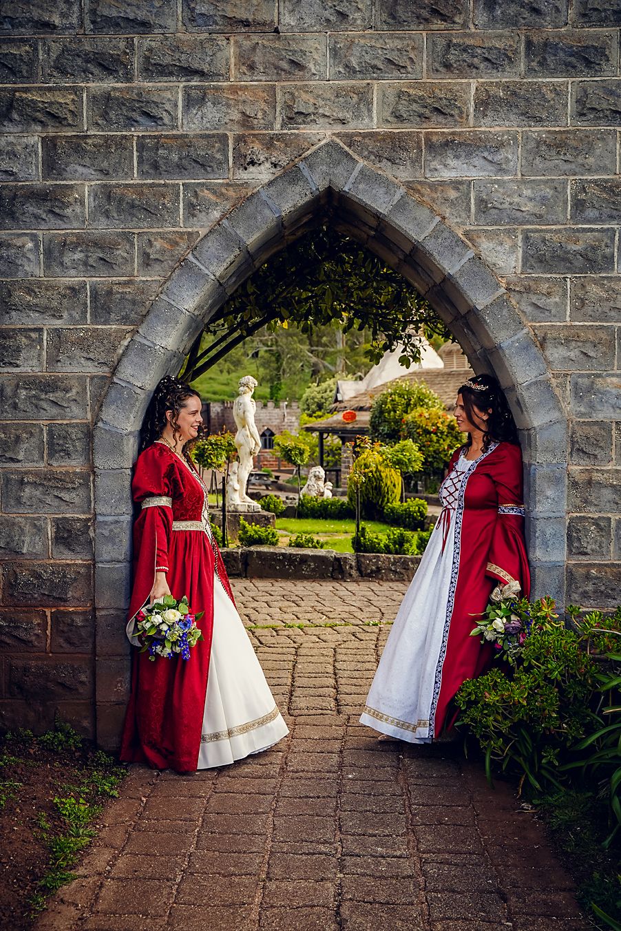Two people in medieval-style red and white dresses stand in a stone archway entrance to a garden path.