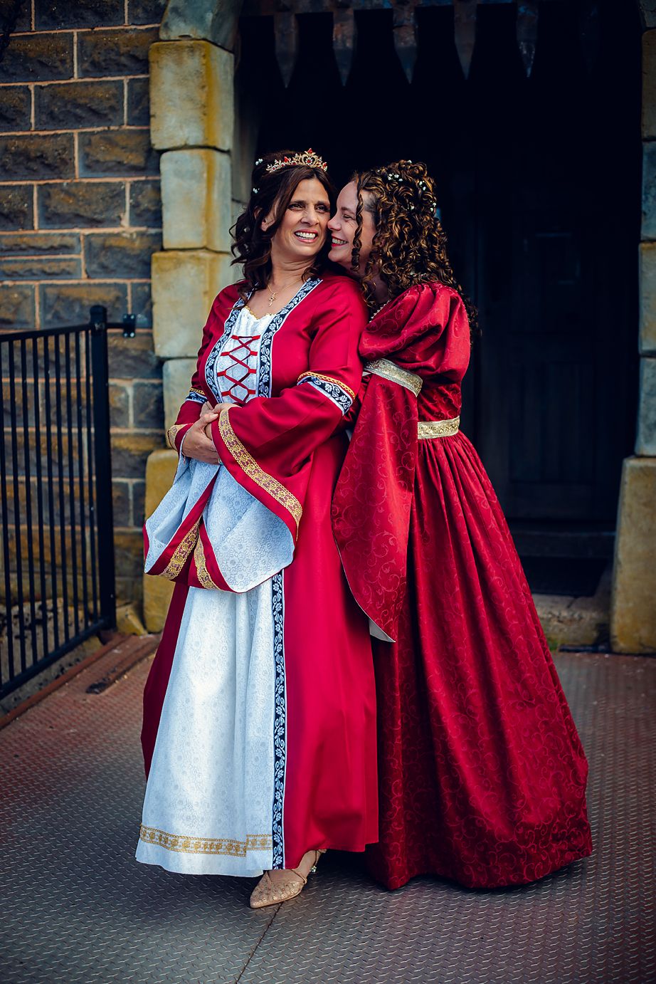 Two people in medieval costumes, one in white and one in red velvet, pose together against a stone wall.