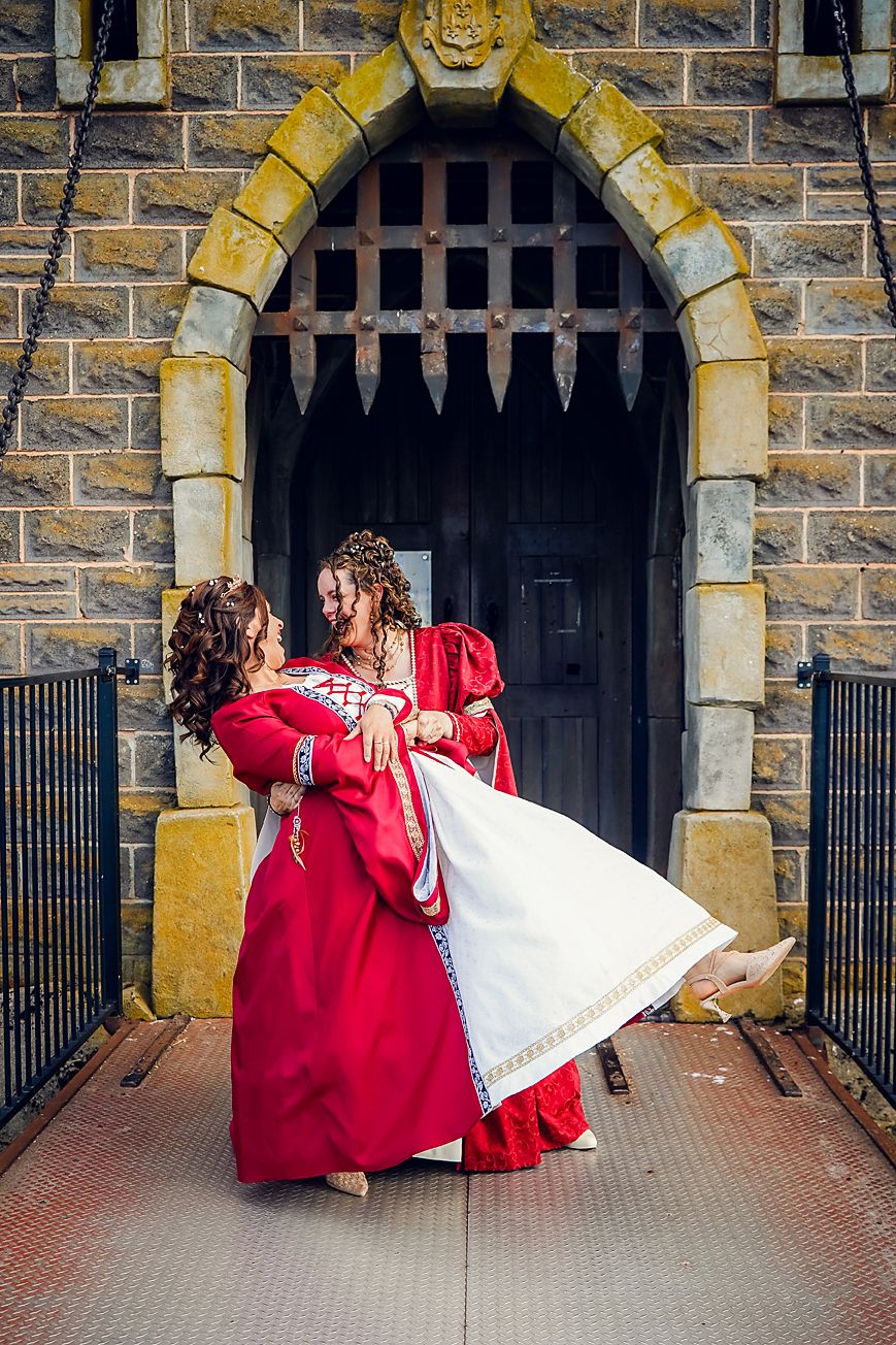 Two people in red medieval style dresses embrace in front of a stone archway with gothic architectural details.