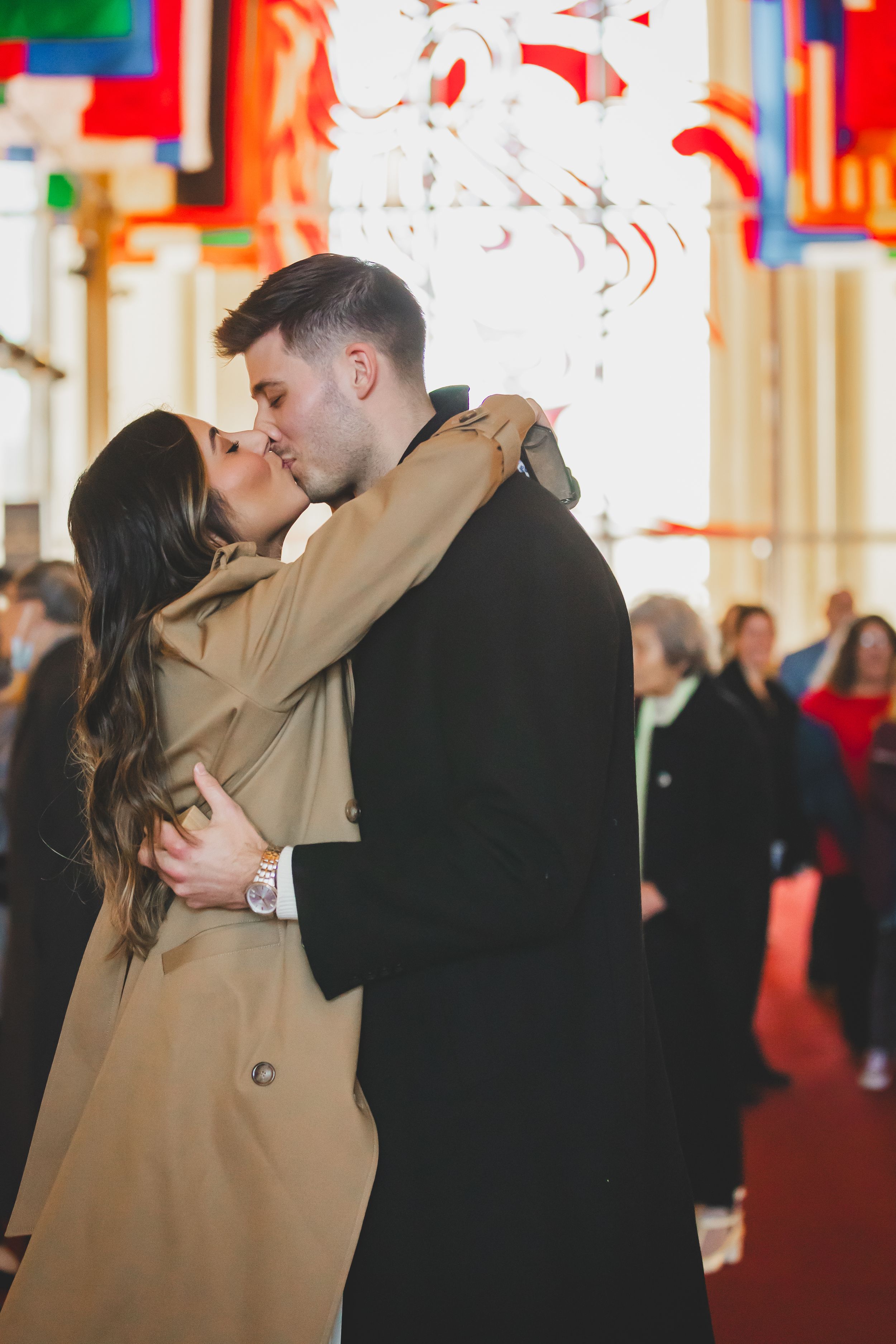 A couple in formal attire share a romantic kiss at an indoor venue with colorful decorations in the background.