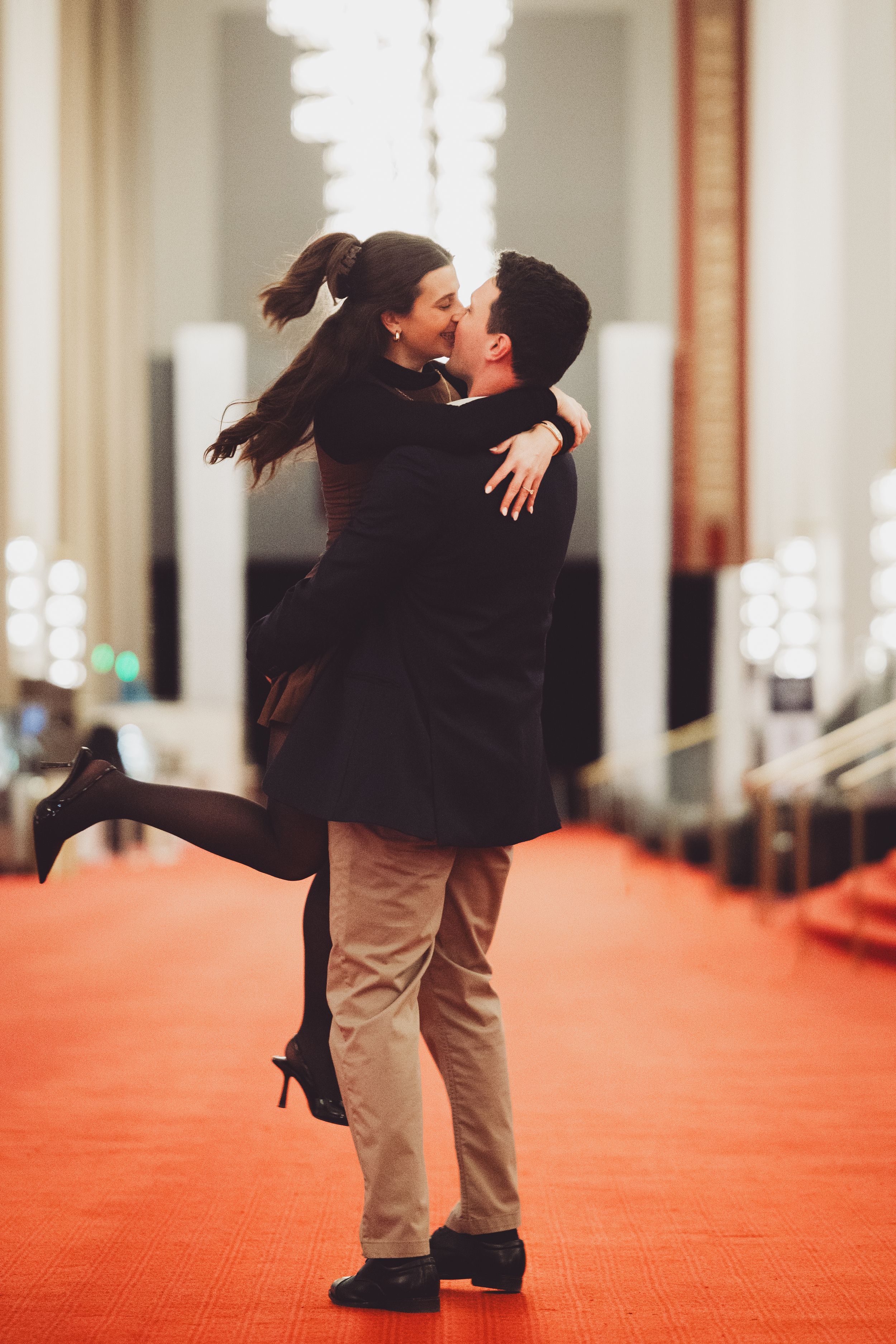 Two people share a romantic embrace on a bright red carpet in a hallway with illuminated white walls of the John F. Kennedy Center in DC