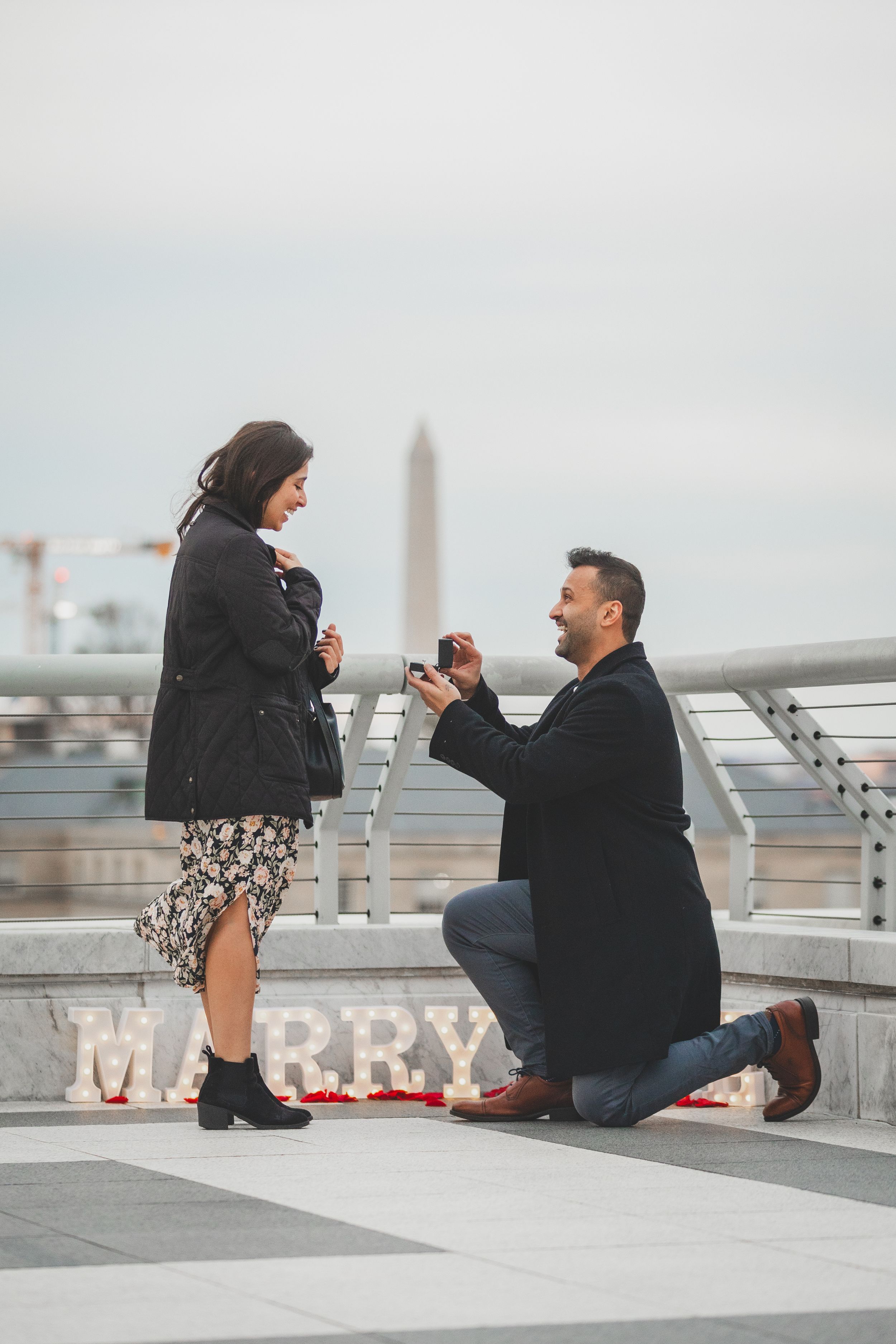 A romantic marriage proposal on a rooftop deck  of the Kennedy Center with 'MARRY' letters and rose petals scattered on the ground.