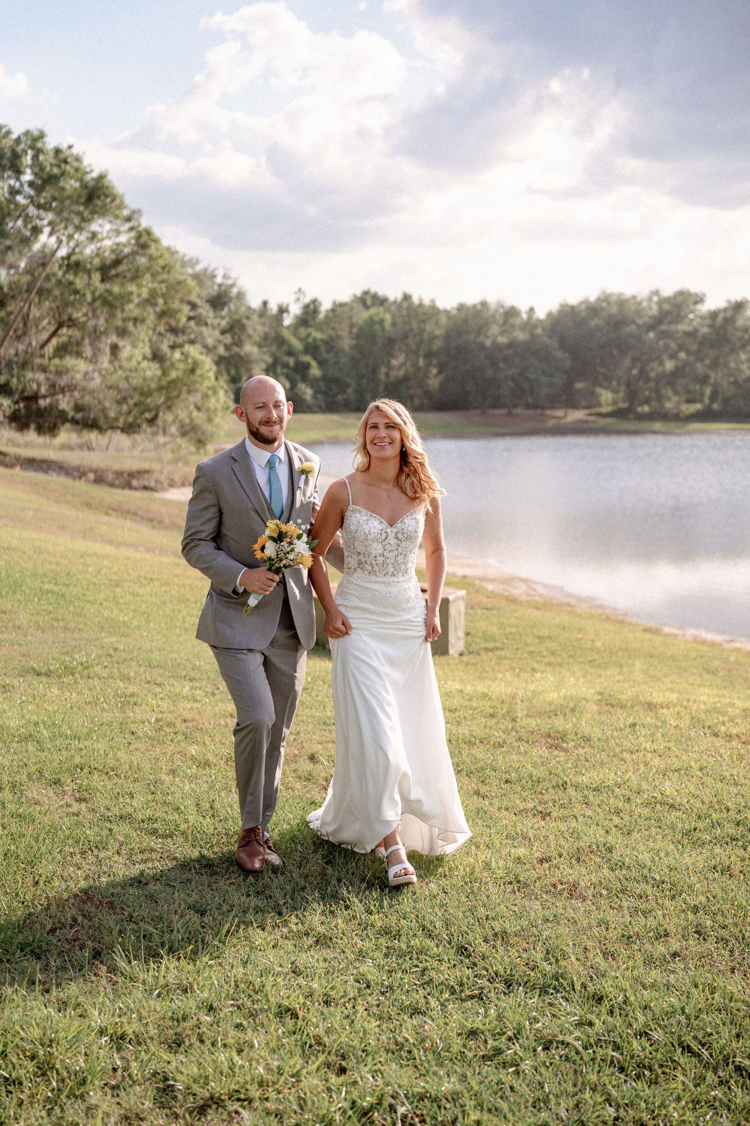 A couple in wedding attire walks together on a grassy hill overlooking a serene lake on their special day.