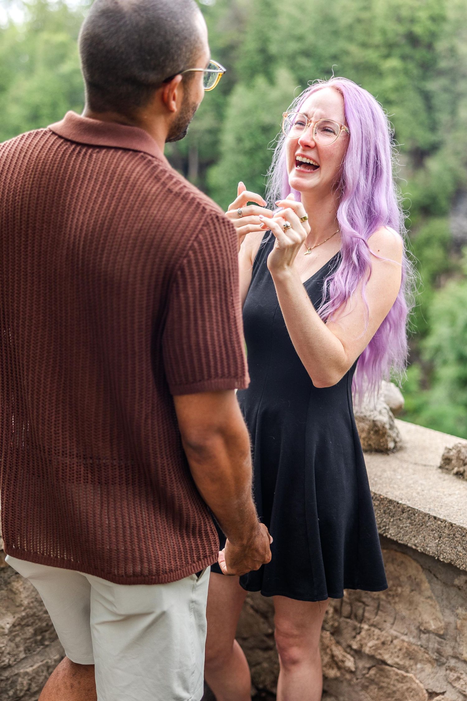 A couple shares a joyful moment together on a scenic overlook, with one person wearing a purple hairstyle and black dress.