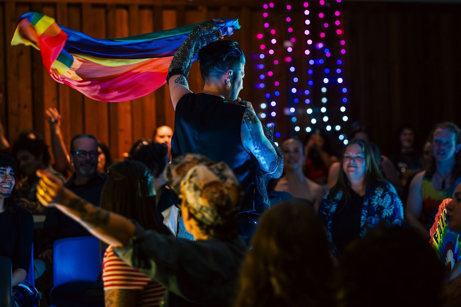 A crowd dances at a nightclub under colorful lights with rainbow flags and decorative lighting creating a festive atmosphere.