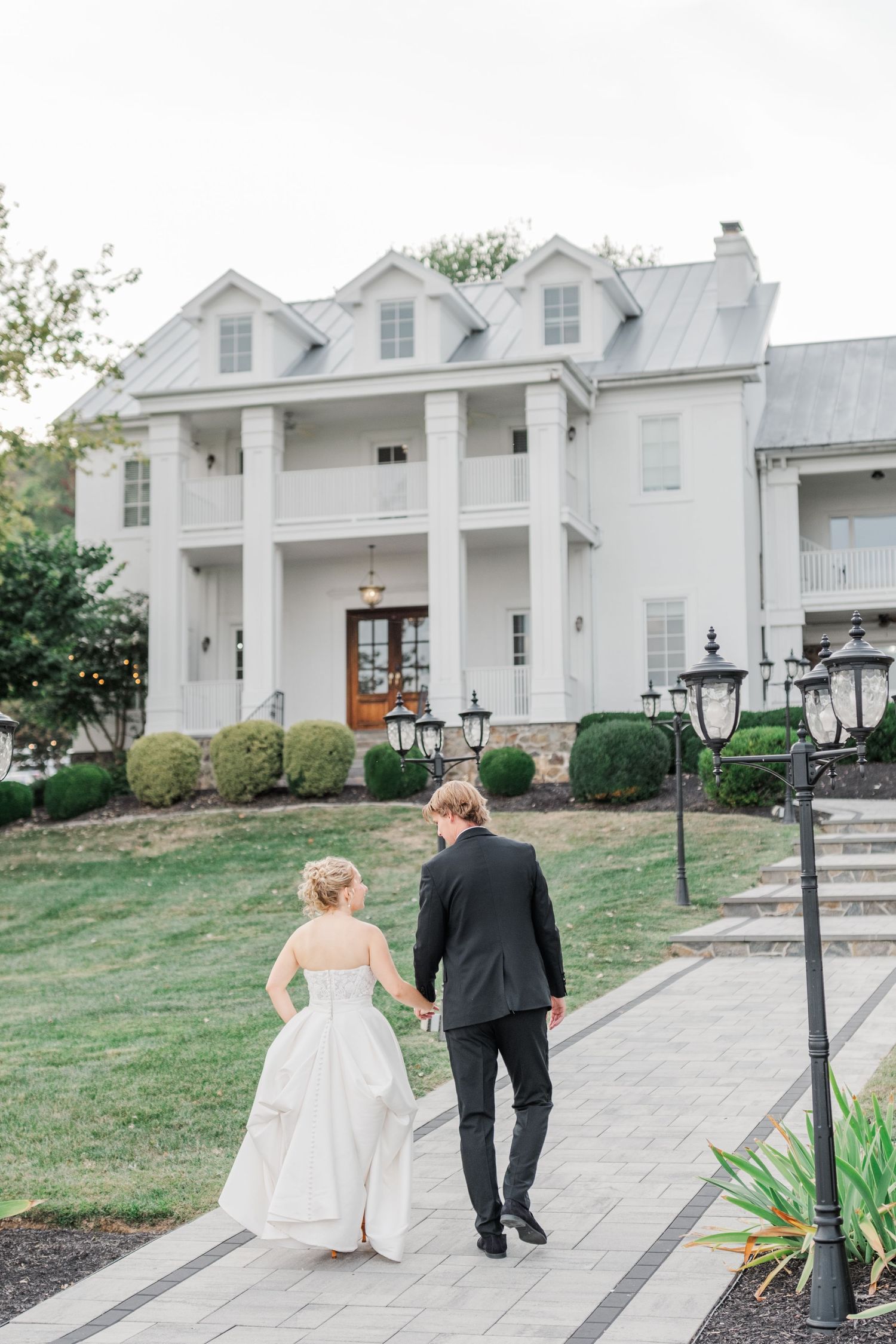 A couple walks along a stone pathway outside a white colonial-style building with dormer windows.