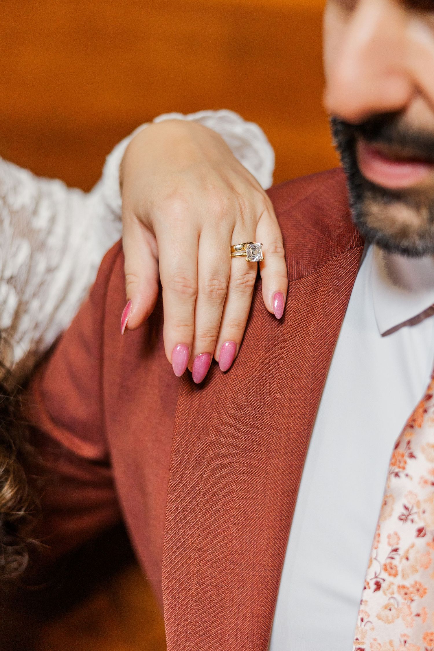 Close-up view of hands with red nail polish and engagement ring resting on a rust-colored fabric surface.
