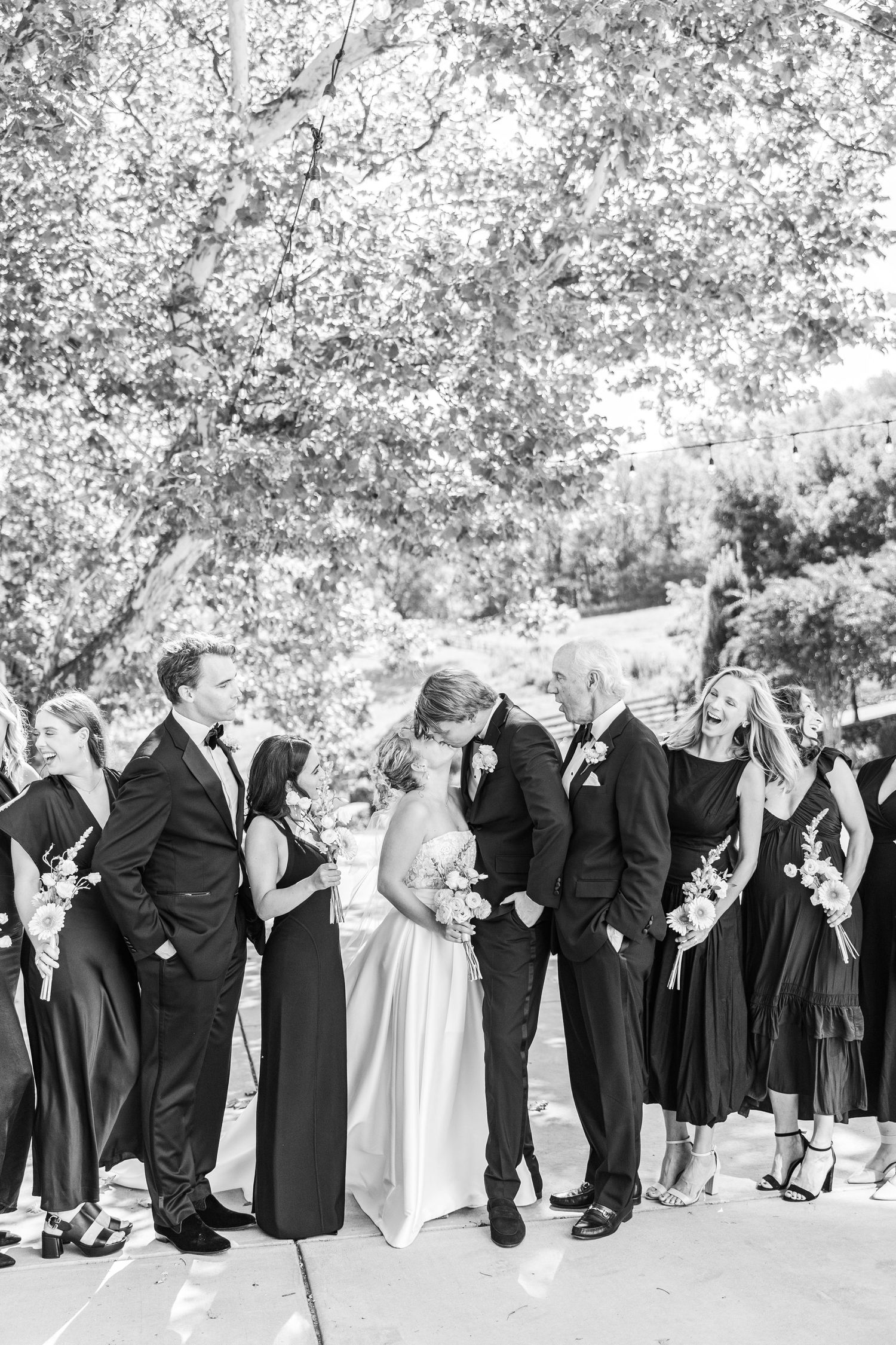 Black and white photo of wedding party members lined up outdoors under trees.
