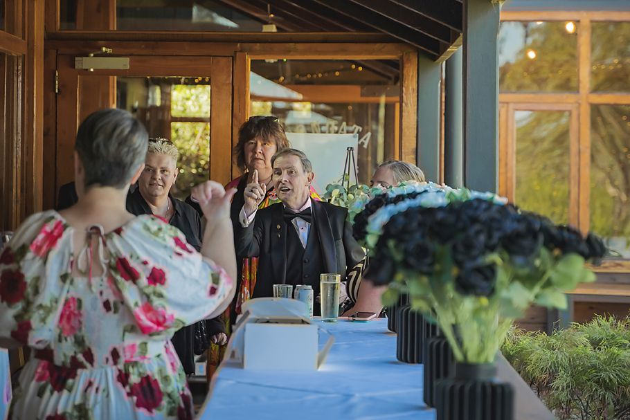 Wedding guests gather for an indoor reception celebration at a rustic venue with large windows and wooden beams.