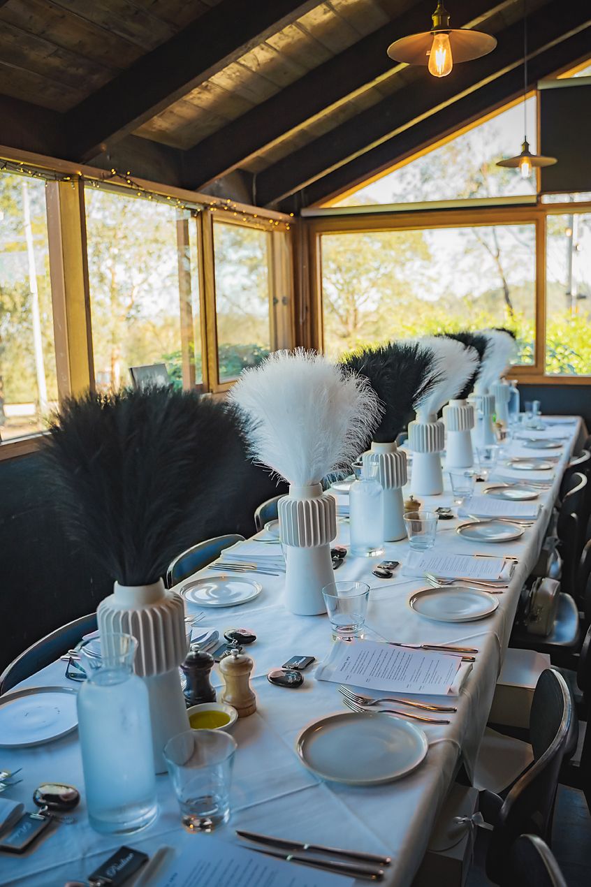 Elegant table setting with white plates and feather centerpieces in a sunlit dining room with panoramic windows.