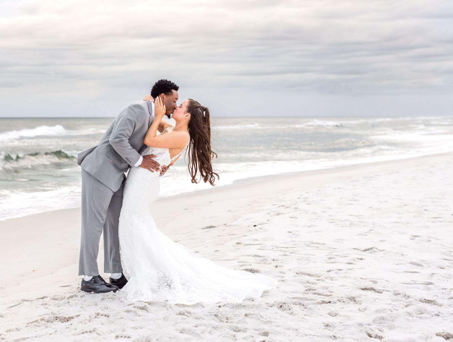 A romantic beach wedding moment with a couple embracing on white sand as ocean waves roll in under cloudy skies.