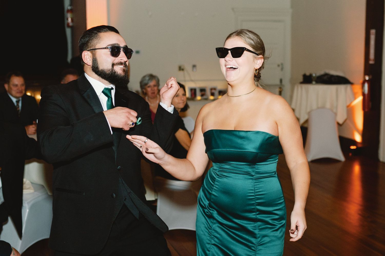 A couple in sunglasses dancing energetically at a reception, with one wearing an emerald green dress.