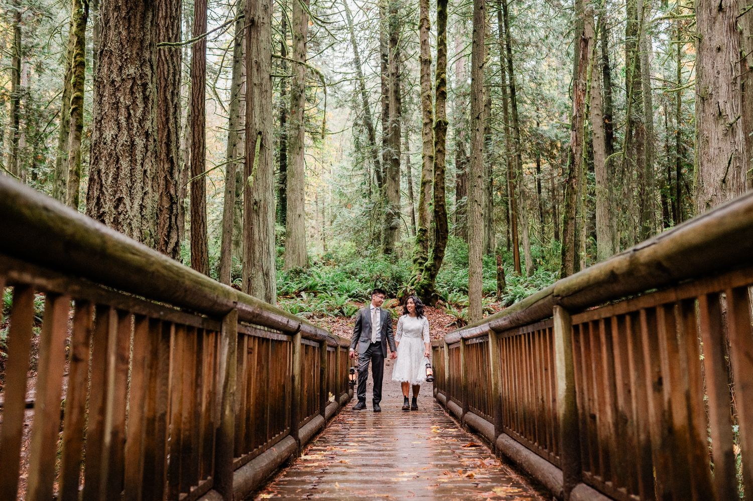 bride and groom walking along a wooden bridge through a lush forest of tall evergreen trees.