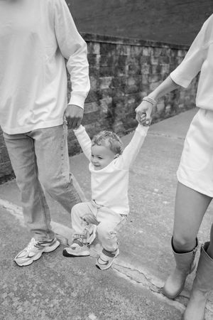 Black and white photo of toddler holding parents' hands while walking on concrete path.