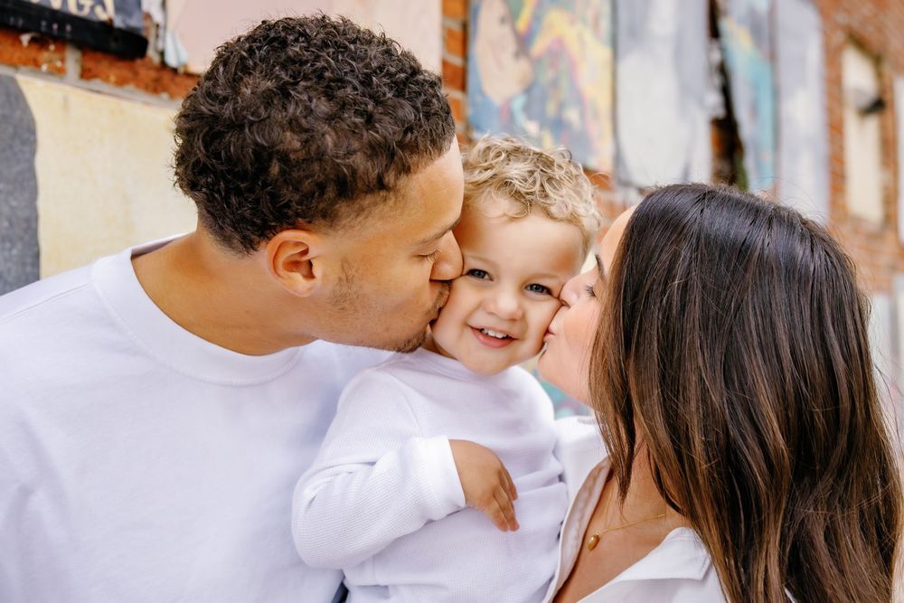 A family shares an intimate moment of kisses and laughter against a rustic wall backdrop.