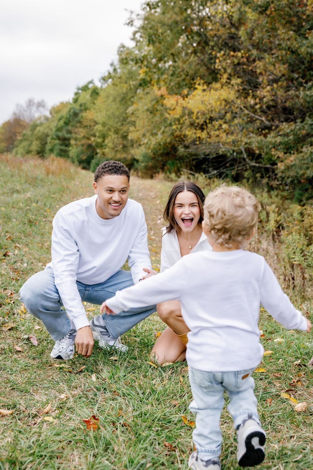 Parents crouch down with excitement as toddler runs towards them in autumn field setting.