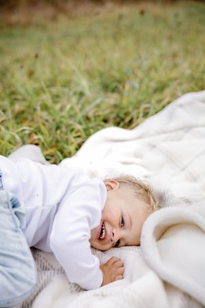 Laughing toddler lies on white blanket in grass during outdoor photoshoot.