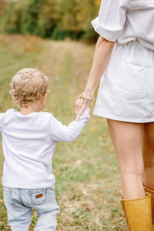 Small child holds parent's hand while walking outdoors, parent wearing white outfit and brown boots.