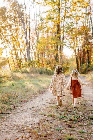 Two children in dresses walk hand in hand down autumn trail surrounded by golden trees.