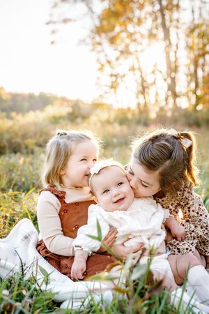 Three children share sweet embraces while sitting on blanket in sunlit autumn field.