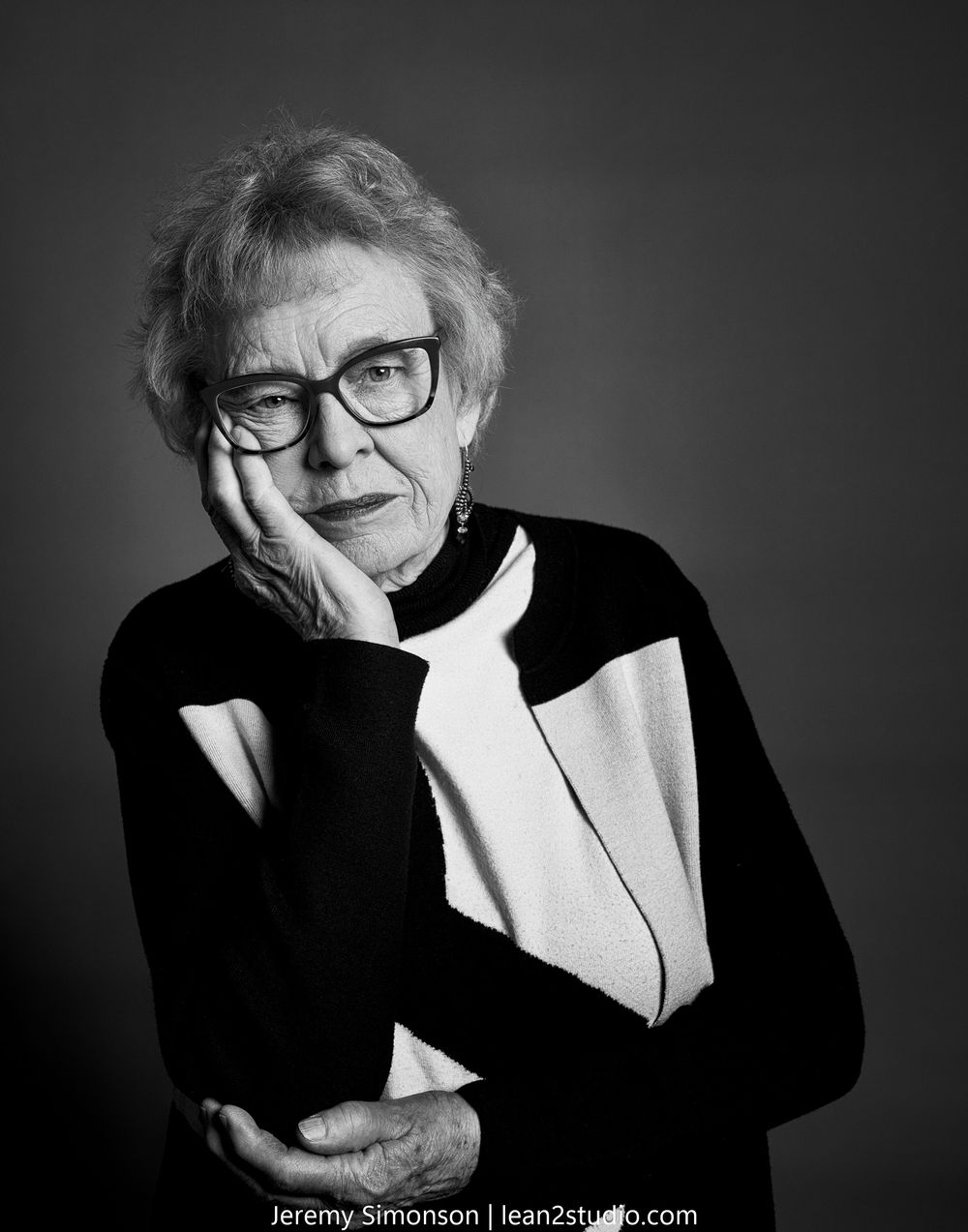 Black and white portrait of someone wearing glasses and a black sweater with white accent against dark background.