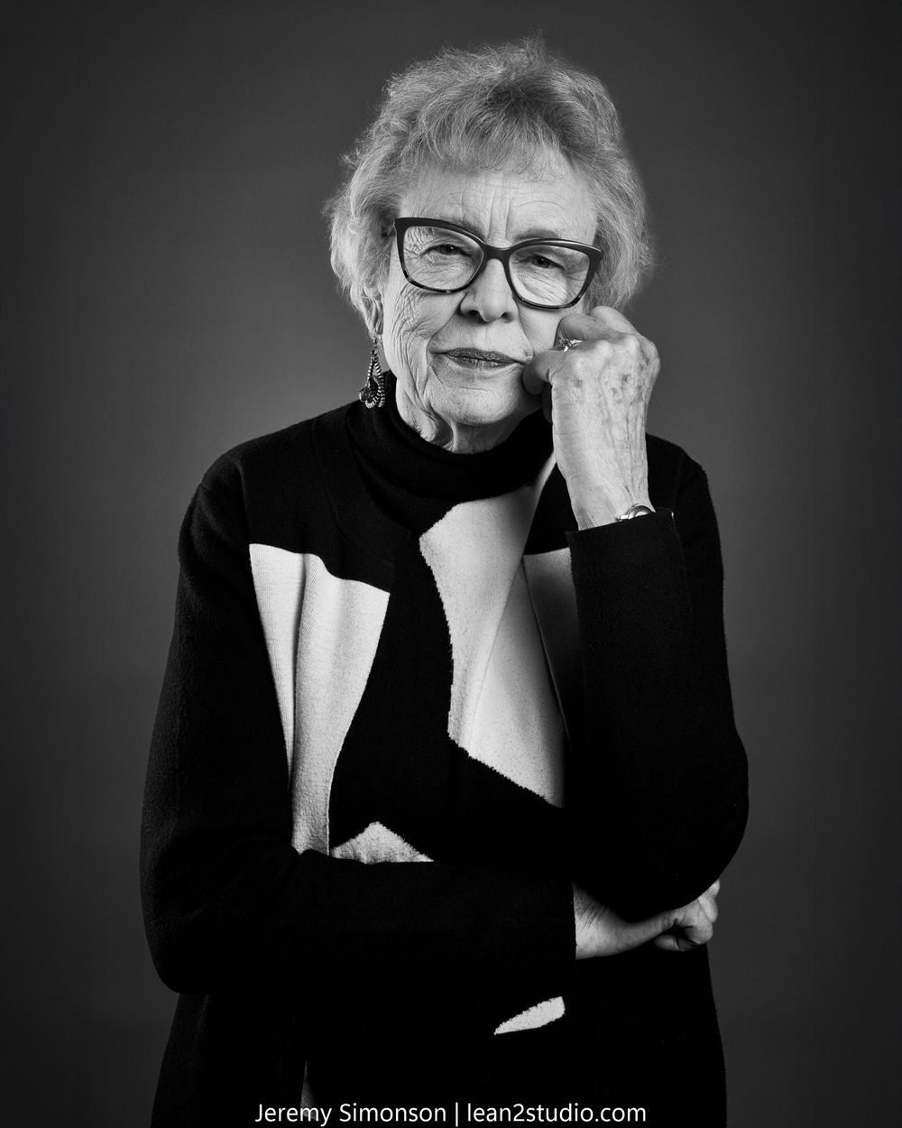 Black and white portrait of an elderly person in glasses and black turtleneck with hand thoughtfully posed near face.