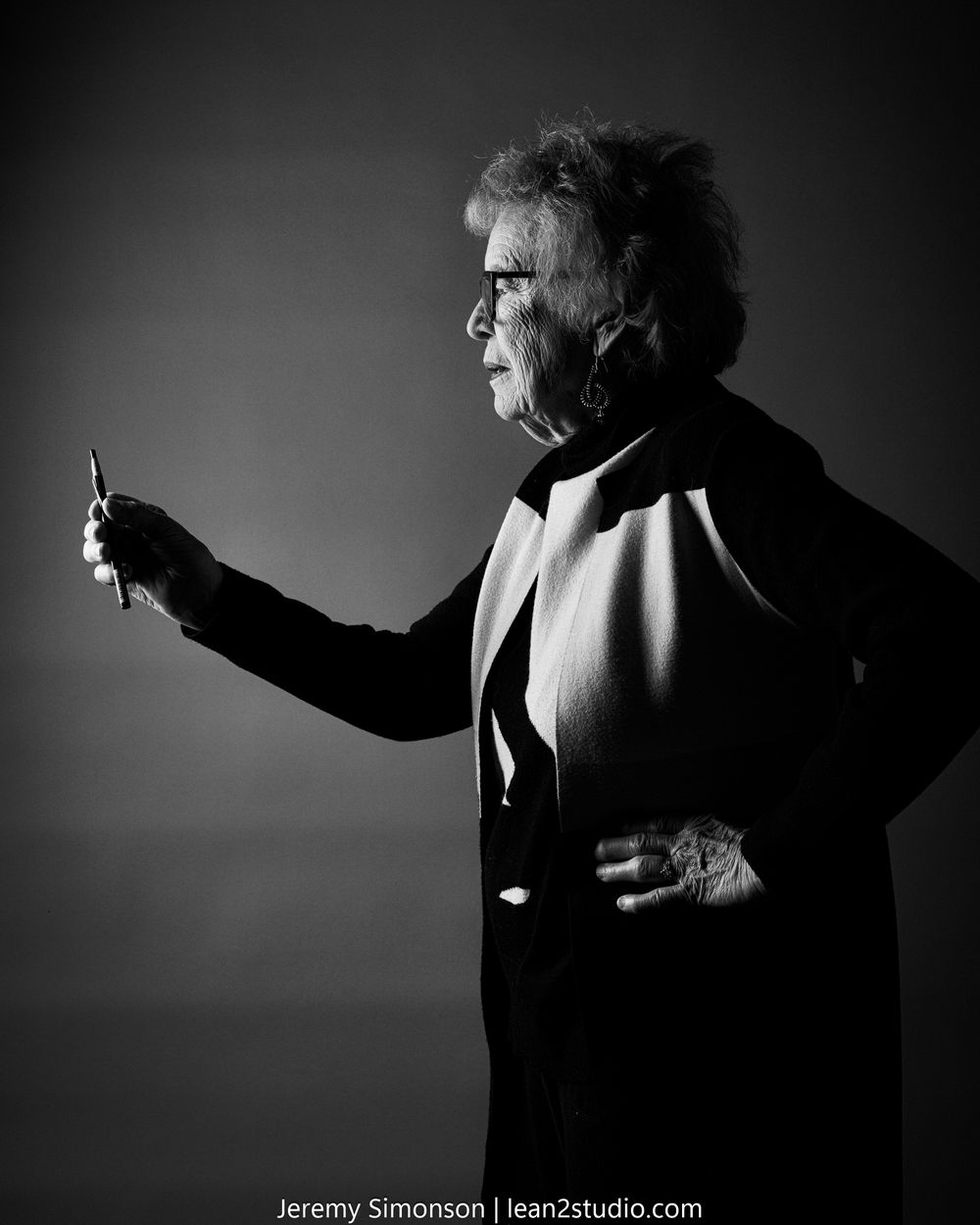 Dramatic black and white profile portrait of a person in glasses and suit holding up an object against dark background.