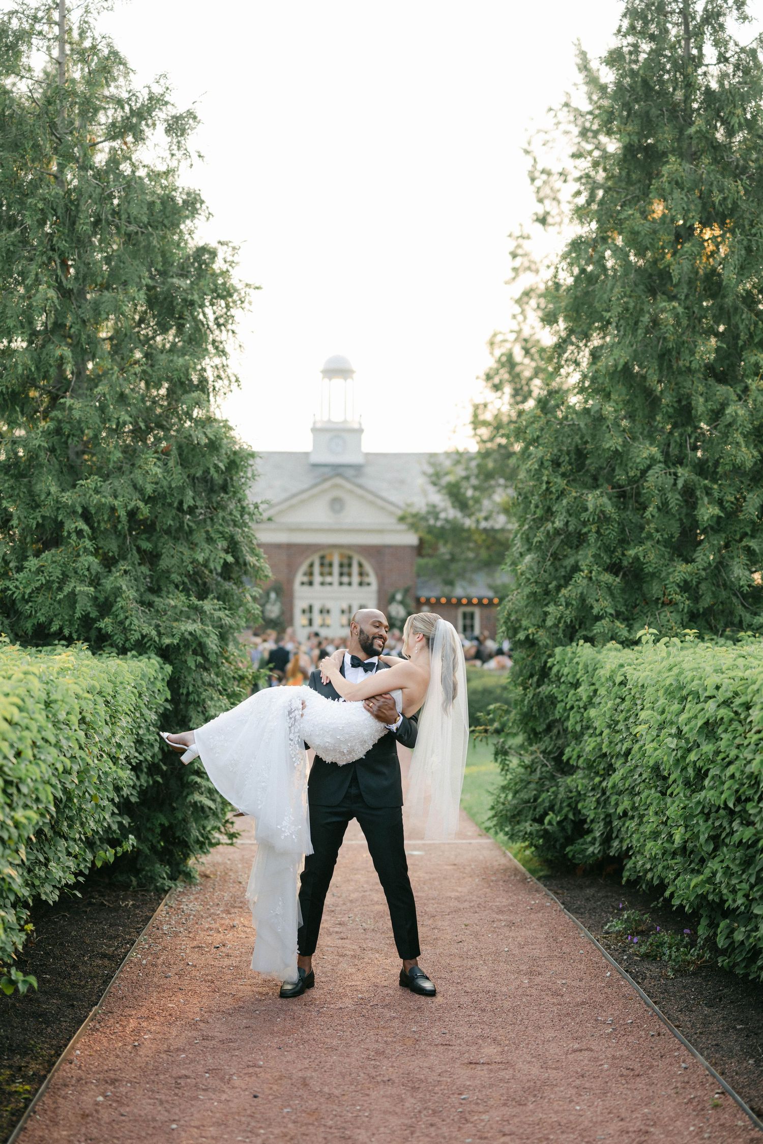 A couple in wedding attire dips playfully on a garden path lined with lush green bushes and trees. - Elawa Farm