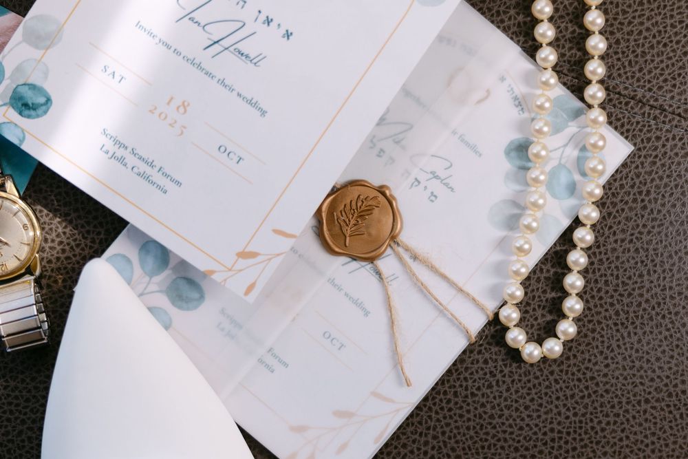 Elegant wedding invitations with pearl necklace, wax seal, and eucalyptus leaves arranged on dark textured surface.