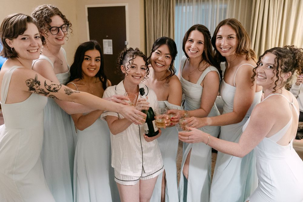 A group in white dresses and robes toasts with champagne bottles during a wedding preparation celebration.