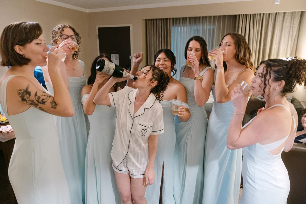 Bridesmaids in light blue dresses stand in a row drinking from champagne glasses during wedding preparations.