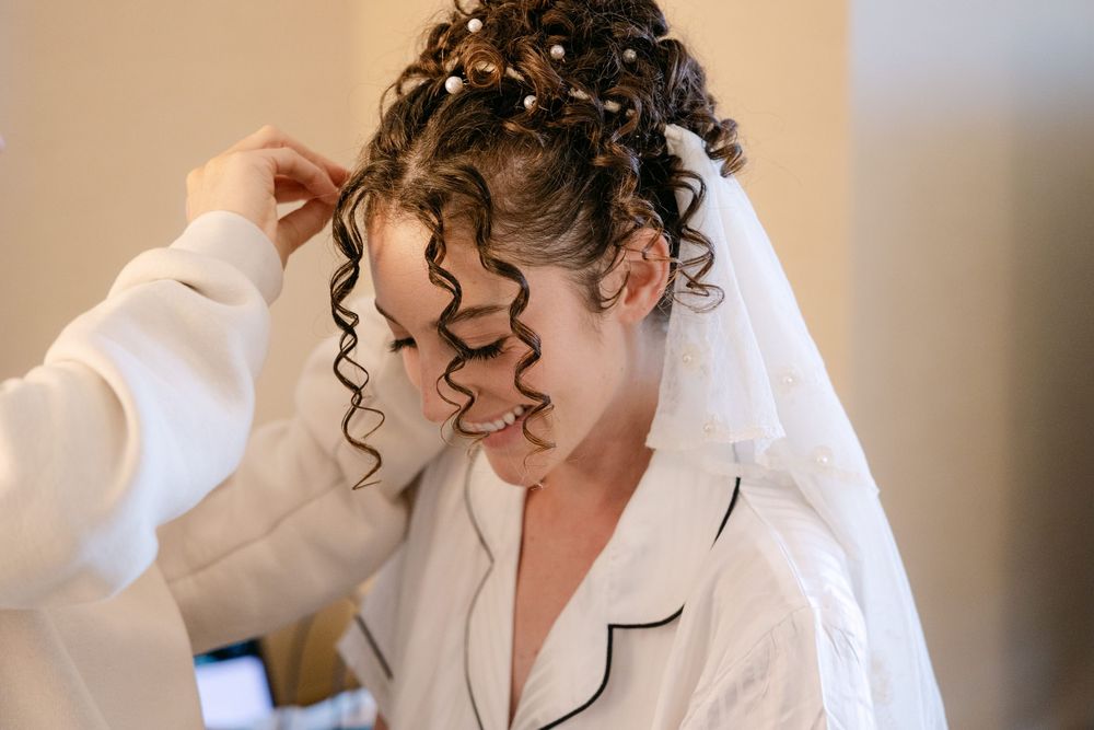 A person getting their curly hair styled while wearing a white silk robe with black trim during wedding preparations.